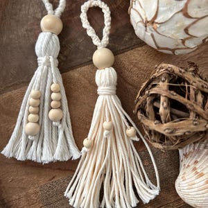May include: Two off-white macrame tassels with wooden beads. Each tassel has a loop for hanging and a cluster of wooden beads. The tassels are displayed with decorative natural elements, including a moss ball and a shell.