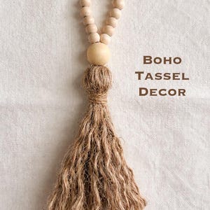 May include: A natural-toned boho tassel decor piece. It features a loop of small wooden beads, a large wooden bead, and a long, textured jute tassel. The words "BOHO TASSEL DECOR" are printed in brown.