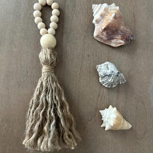 May include: A decorative tassel made of natural jute twine featuring a loop of light-colored wooden beads at the top, accompanied by three distinct seashells of varying sizes and brown, white, and grey tones arranged to the right of the tassel.