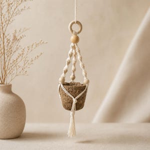 May include: A hanging plant holder made of off-white macrame cord featuring twisted patterns, light-colored wooden beads, and a wooden ring at the top. The holder contains a small, textured, brown, cone-shaped pot with a tassel at the bottom.