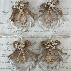 May include: Four handmade angel ornaments. Each angel features a wooden bead body, a jute bow, and a halo of white rope. The wings are made of jute and the head is adorned with small white flowers. Each ornament hangs from a white string.