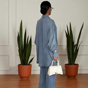 May include: A denim blue, long-sleeved shirt and matching wide-leg pants. The shirt has a button-down front and embellished cuffs. A white handbag with a silver clasp is held in the hand. The outfit is paired with a pair of glasses and earrings.