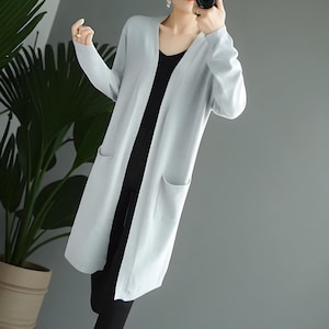 Op de afbeelding: Een lichtblauwe lange cardigan met een V-hals en twee voorzakken. De cardigan is knielang en lijkt gemaakt van een zacht gebreid materiaal. De cardigan wordt over een zwarte outfit gedragen.