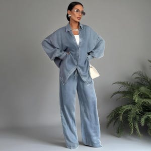May include: A light blue denim outfit, consisting of a button-down shirt and wide-leg pants. The shirt has a classic collar and long sleeves. The pants have a high waist and a relaxed fit. A white purse is worn over the shoulder. The outfit is a casual, yet stylish ensemble.