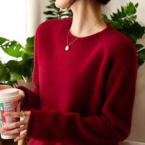May include: A solid red crew neck sweater with long sleeves. A gold necklace with a circular pendant is worn. The person is holding a white coffee cup with a red and green design. The background includes green leafy plants.