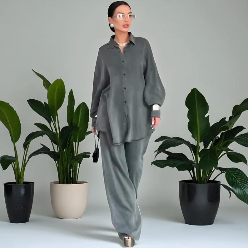 May include: A two-piece set of gray-green clothing. The top is a button-down shirt with long sleeves and a relaxed fit. The pants are wide-legged. The outfit is worn with silver accessories and gold platform sandals. Potted plants are in the background.