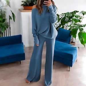 Op de afbeelding: Een tweedelige gebreide set in een gedempte blauwe tint. De set bestaat uit een trui met lange mouwen en blote schouders en een broek met wijde pijpen. De outfit is gemaakt van een zacht, getextureerd materiaal, geschikt voor casual wear. Het ensemble wordt getoond in een goed verlichte binnenomgeving.