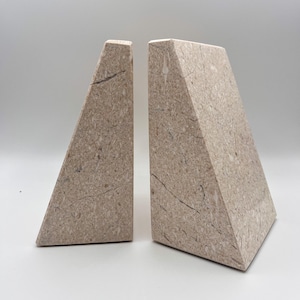 May include: Two beige marble bookends with a triangular prism shape. The bookends have a natural stone texture with dark gray veining. The bookends are designed to stand upright and support books on a shelf.