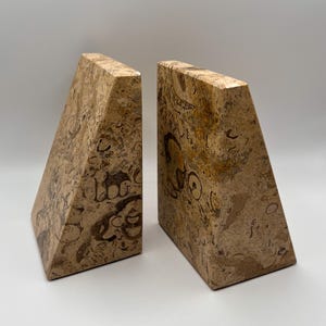 May include: Two beige stone bookends with a triangular shape. The bookends have a natural, patterned surface with brown and tan markings. The bookends are designed to hold books upright on a shelf or desk.