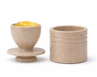 May include: A two-piece butter dish set. The butter dish is beige with a pedestal base and a bowl-shaped top filled with yellow butter. The lid is a cylindrical container with horizontal grooves. The set is made of a stone-like material.