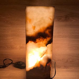 May include: A rectangular onyx lamp with a warm, glowing interior. The lamp features a mix of light beige and dark brown tones, with a black power cord and foot switch. The lamp is set against a wooden background.