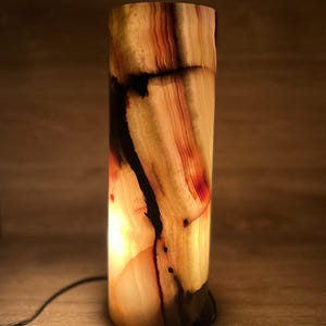 May include: A cylindrical lamp made of translucent stone, showcasing a warm, glowing interior. The stone features bands of beige, red, and black, creating a natural, textured pattern. A black power cord extends from the base. This decorative lamp is ideal for home decor.