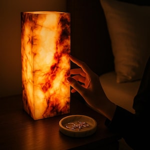 Puede incluir: Una lámpara rectangular de ónix con un interior cálido y brillante. La superficie de la lámpara muestra una mezcla de vetas naranjas y marrones. Una pequeña bandeja redonda de mármol con joyas está cerca.