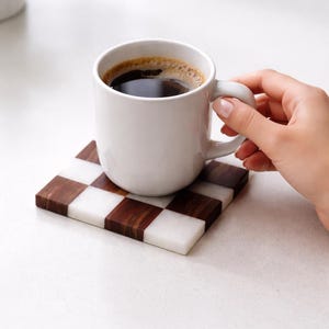 Puede incluir: Una taza blanca llena de caf&eacute; oscuro descansa sobre un posavasos a cuadros. El posavasos presenta un patr&oacute;n de cuadrados marrones y blancos alternados. La taza es sostenida por una mano, sugiriendo una pausa para el caf&eacute;.