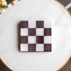 Puede incluir: Un posavasos cuadrado con dise&ntilde;o de tablero de ajedrez con cuadrados marrones oscuros y blancos alternos. El posavasos est&aacute; hecho de madera y m&aacute;rmol. El posavasos est&aacute; sobre una superficie blanca con un borde dorado.