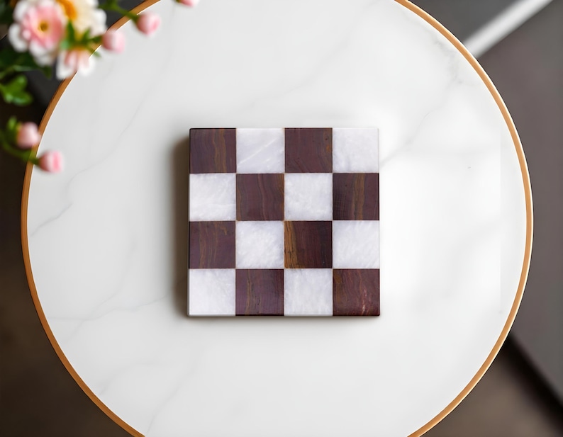 Puede incluir: Un posavasos cuadrado con dise&ntilde;o de tablero de ajedrez con cuadrados marrones oscuros y blancos alternos. El posavasos est&aacute; hecho de madera y m&aacute;rmol. El posavasos est&aacute; sobre una superficie blanca con un borde dorado.