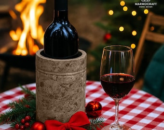 May include: A bottle of red wine in a stone wine chiller, with a glass of red wine on a red and white checkered tablecloth. A red bow and ornaments decorate the table. The background includes a fire and Christmas lights.