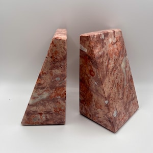 Puede incluir: Dos sujetalibros de mármol rojo y blanco. Los sujetalibros tienen un diseño geométrico con forma triangular. El mármol presenta un patrón natural de vetas rojas, blancas y grises. Diseñados para mantener los libros en posición vertical.