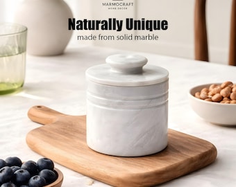 May include: A white marble container with a lid, sitting on a wooden cutting board. The container has a cylindrical shape with horizontal grooves. The text "Naturally Unique made from solid marble" is displayed above the container. A bowl of blueberries is in the foreground.