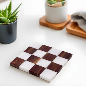Puede incluir: Posavasos cuadrado con un dise&ntilde;o de tablero de ajedrez. Cuadrados alternos de m&aacute;rmol blanco y madera oscura crean un contraste llamativo. El posavasos est&aacute; sobre una superficie blanca, con una planta en maceta y otros elementos decorativos en el fondo.