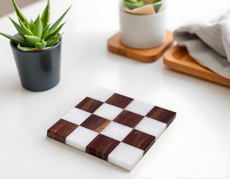 Puede incluir: Posavasos cuadrado con un dise&ntilde;o de tablero de ajedrez. Cuadrados alternos de m&aacute;rmol blanco y madera oscura crean un contraste llamativo. El posavasos est&aacute; sobre una superficie blanca, con una planta en maceta y otros elementos decorativos en el fondo.