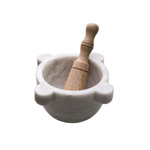 May include: A white marble mortar and pestle set. The mortar has a rounded bowl shape with four small, rounded protrusions. A wooden pestle rests inside the mortar. The pestle is light brown and has a rounded handle.