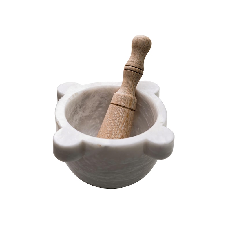 May include: A white marble mortar and pestle set. The mortar has a rounded bowl shape with four small, rounded protrusions. A wooden pestle rests inside the mortar. The pestle is light brown and has a rounded handle.