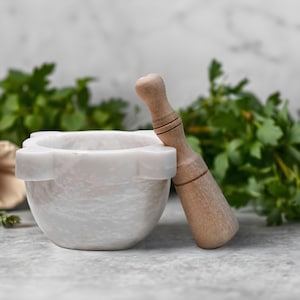 May include: A white marble mortar and pestle set with a wooden pestle, perfect for grinding herbs and spices. The mortar has a rounded bowl shape with a decorative rim. Fresh green herbs and a red tomato are in the background.