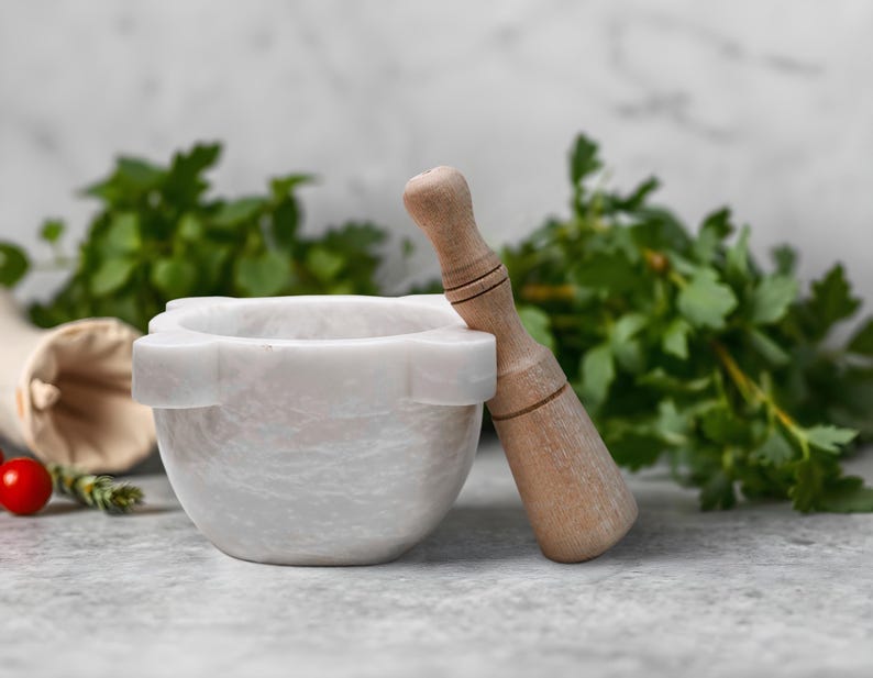 May include: A white marble mortar and pestle set with a wooden pestle, perfect for grinding herbs and spices. The mortar has a rounded bowl shape with a decorative rim. Fresh green herbs and a red tomato are in the background.