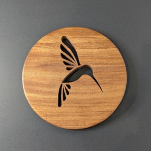 May include: A circular wooden coaster featuring a hummingbird cut-out. The coaster is made of light brown wood, with a detailed silhouette of a hummingbird in flight. The background is a solid dark grey.