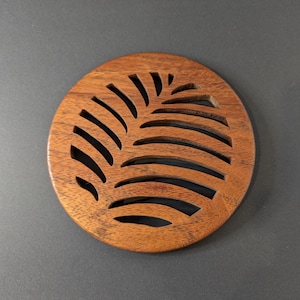 May include: A round, wooden coaster with a leaf-like design cut into the surface. The coaster is made of a dark brown wood and has a smooth finish. The design features curved lines that create a pattern.