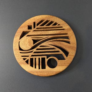 Può includere: Un sottobicchiere rotondo in legno con un design geometrico. Il sottobicchiere presenta una serie di forme ritagliate, tra cui linee, curve e cerchi. Il legno ha un tono caldo e naturale e il sottobicchiere è posto su uno sfondo grigio scuro.