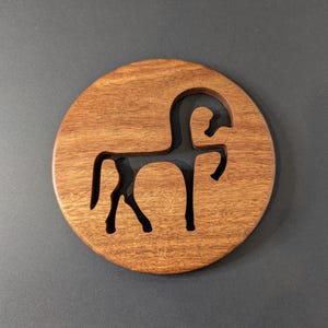 May include: A round wooden coaster with a carved horse silhouette. The coaster is made of a warm-toned wood and features a dark horse shape cut out in the center. The design is simple and elegant, suitable for various decor styles.