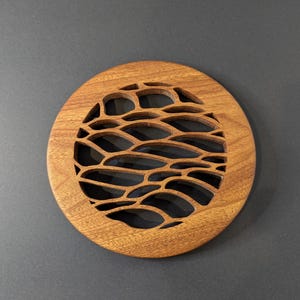 May include: A round, wooden coaster with a decorative, wave-like cutout pattern. The coaster is made of light brown wood and sits on a dark gray surface. The coaster's design is a series of interconnected, organic shapes.