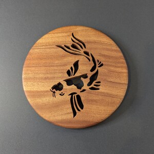 May include: Round wooden coaster with a carved koi fish design. The coaster is made of a light brown wood and features a black and white koi fish silhouette. The coaster is a decorative item for home decor.