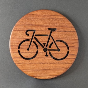 May include: A round wooden coaster with a bicycle cutout design. The coaster is made of a light brown wood with visible grain and a dark gray background. The bicycle is a simple outline in black.