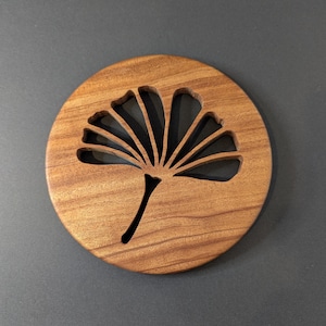 May include: A round wooden coaster with a cut-out ginkgo leaf design. The coaster is made of a light brown wood with a visible grain pattern. The ginkgo leaf design is cut out of the wood, creating a negative space.