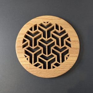 May include: Circular wooden coaster with a geometric pattern. The coaster is made of light-colored wood and features a repeating design of interlocking shapes. The design is cut out, creating negative space within the coaster's surface.