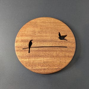 May include: A round wooden coaster with a bird silhouette perched on a line, and another bird in flight. The coaster is made of a light brown wood with a visible grain pattern. The background is a solid dark gray.
