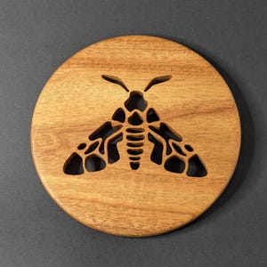 May include: A round, wooden coaster with a carved-out moth design. The coaster is made of light brown wood and has a smooth finish. The moth design is detailed, with visible wings, body segments, and antennae. The coaster is a decorative item.