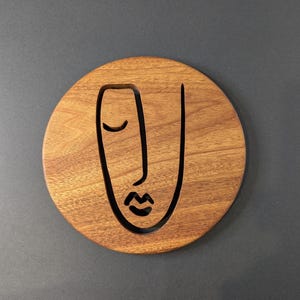 May include: A round wooden coaster with a carved face design. The coaster is made of a light brown wood and features a simple, minimalist face with a closed eye, nose, and lips. The coaster is a decorative item.