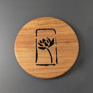May include: A round wooden coaster with a carved floral design. The coaster is made of light brown wood and features a dark floral silhouette within a rectangular frame. The coaster is designed to protect surfaces from drink condensation.