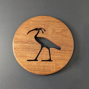 May include: A round wooden coaster with a dark brown finish. A black silhouette of a bird is cut out of the wood. The coaster is on a gray surface.