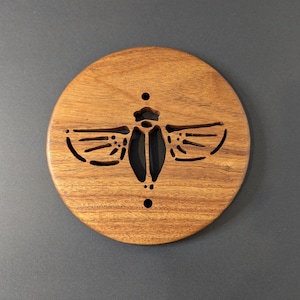 May include: A round wooden coaster with a laser-cut insect design. The coaster is made of light brown wood and features a detailed silhouette of a beetle. The insect's body is dark, and the wings are cut out, creating a decorative accent.