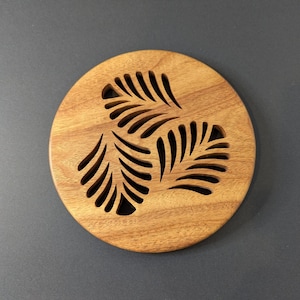 May include: A round wooden coaster with a natural finish. The coaster features three leaf-like cutouts arranged in a triangular pattern. The coaster is approximately 4 inches in diameter.