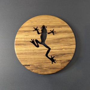 May include: A round wooden coaster with a dark frog silhouette cut out of the center. The coaster has a natural wood grain pattern and a dark frog design. The frog is in a leaping pose.