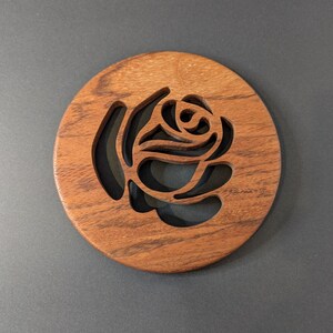 May include: A round wooden coaster with a rose design cut out of the center. The coaster is made of a dark brown wood with visible grain and sits on a gray surface. The rose design is a negative space.