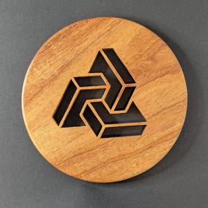 May include: A round wooden coaster with a geometric design. The coaster is made of light brown wood and features a dark, three-dimensional symbol cut out of the center. The symbol is a stylized, interlocking triangle.