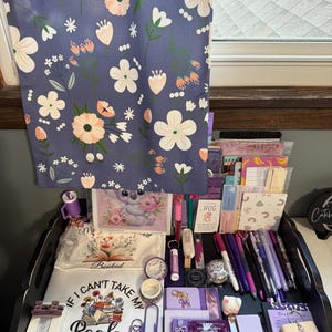 Peut inclure: Un sac fourre-tout bleu &agrave; motif floral blanc et p&ecirc;che est suspendu au-dessus d'un plateau noir rempli de papeterie, incluant des stylos violets, des carnets, une pochette blanche avec le texte "I CAN'T TAKE MY BOOK I'M NOT GOING" et un koala.