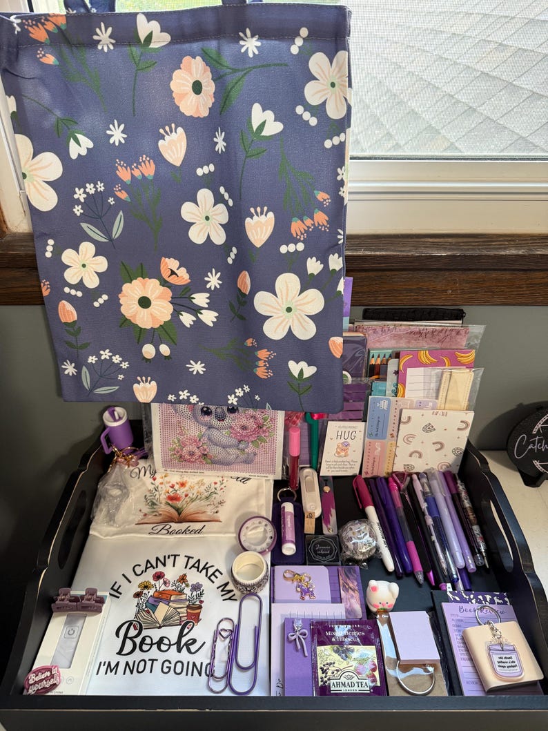 Peut inclure: Un sac fourre-tout bleu &agrave; motif floral blanc et p&ecirc;che est suspendu au-dessus d'un plateau noir rempli de papeterie, incluant des stylos violets, des carnets, une pochette blanche avec le texte "I CAN'T TAKE MY BOOK I'M NOT GOING" et un koala.